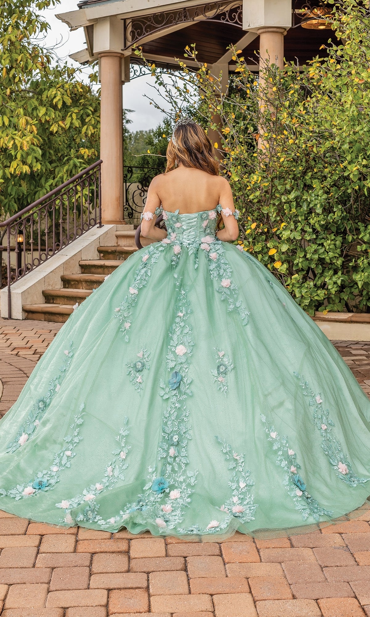 A woman in the Quinceanera Dress 1828 by Dancing Queen, featuring an off-the-shoulder sage green design with floral embellishments, stands outdoors with her back to the camera near stone steps and greenery.